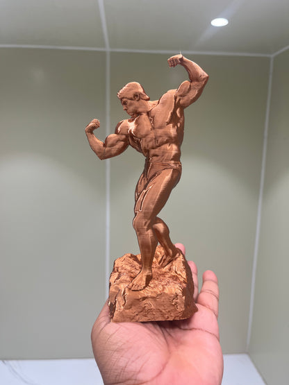 A hand holds the 8” Arnold Gym Champion Sculpture—a bronze-toned statue of a flexing bodybuilder on a textured base—set against a light background, making it the perfect gift for gym lovers.