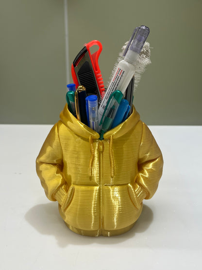 The Mini Hoodie Desk Organizer - 3D Printed Pen Holder, gold-colored and shaped like a hooded jacket, holds pens, markers, a box cutter, and correction tape. It sits on a white surface against a plain background.