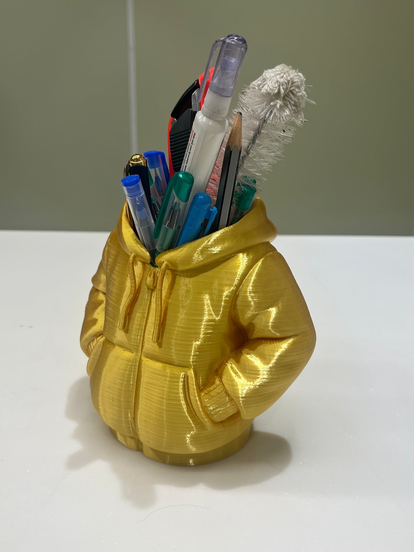 The Mini Hoodie Desk Organizer - 3D Printed Pen Holder, shaped like a yellow hooded jacket and filled with pens and a cleaning brush, sits on a white surface—a creative accessory for any workspace.