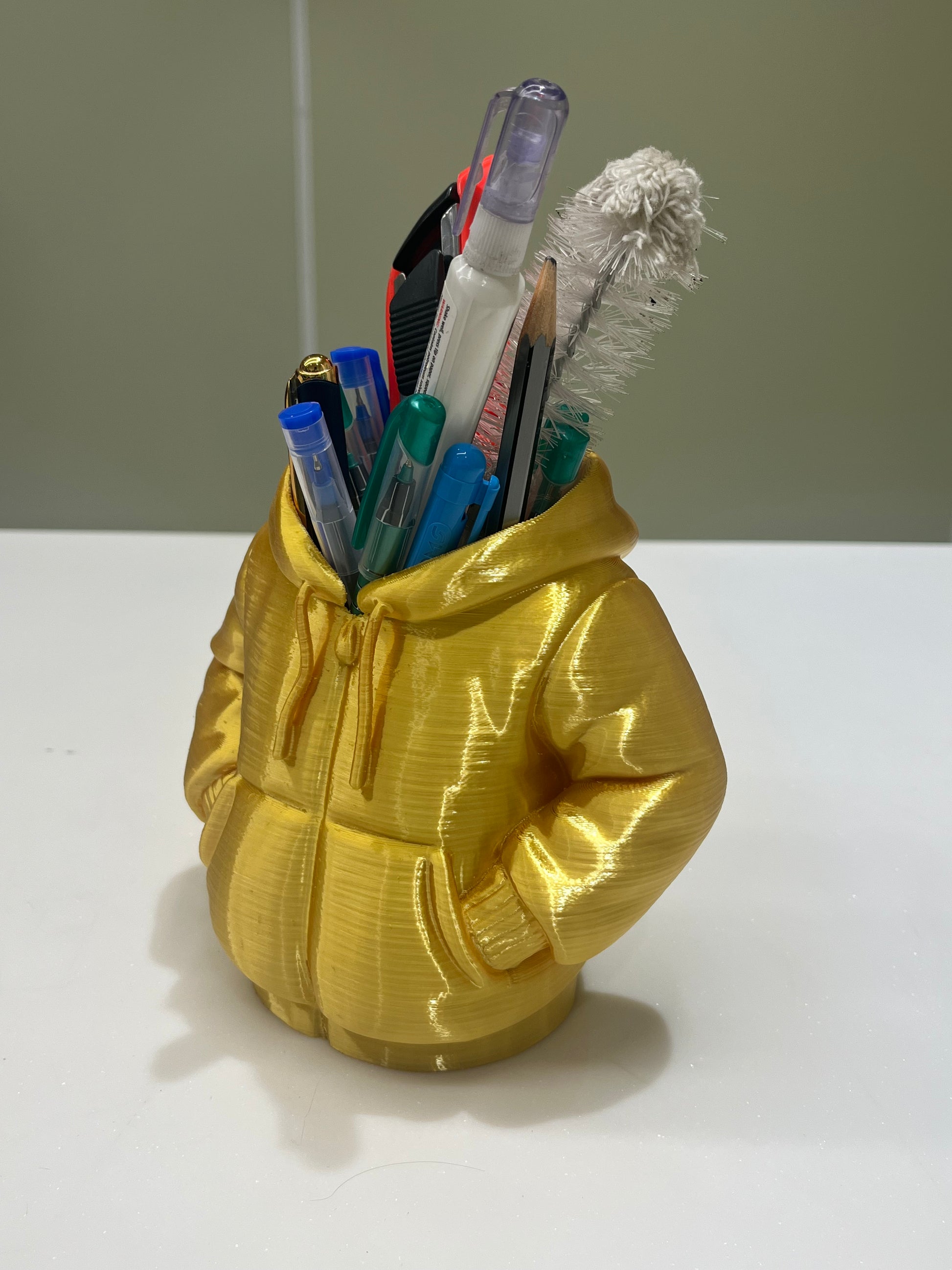 The Mini Hoodie Desk Organizer - 3D Printed Pen Holder, shaped like a yellow hooded jacket and filled with pens and a cleaning brush, sits on a white surface—a creative accessory for any workspace.