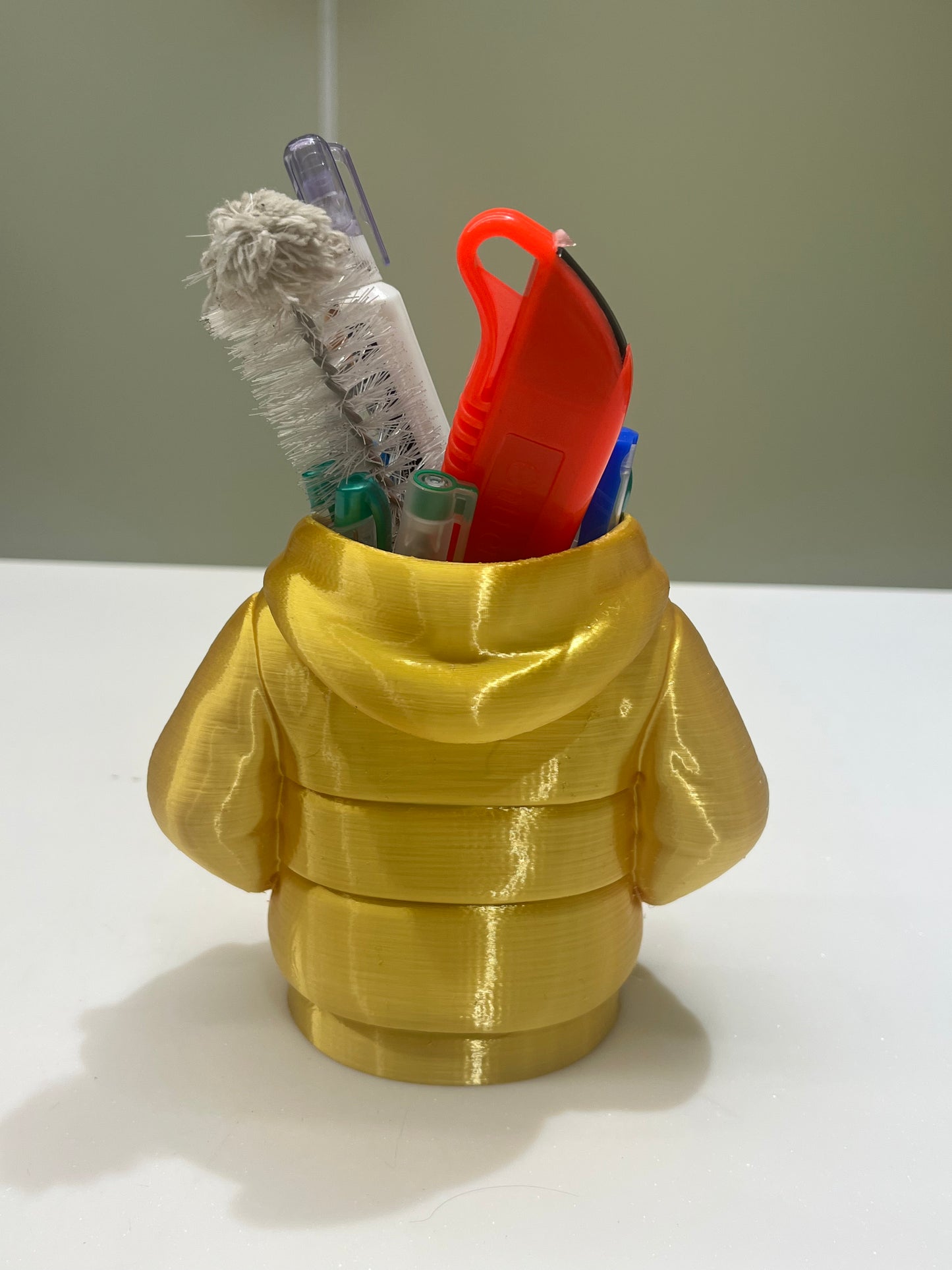 The Mini Hoodie Desk Organizer is a yellow, 3D-printed pen holder shaped like a puffy jacket, storing pens, a utility knife, toothbrush, and cleaning brush—a creative desk accessory for any workspace.