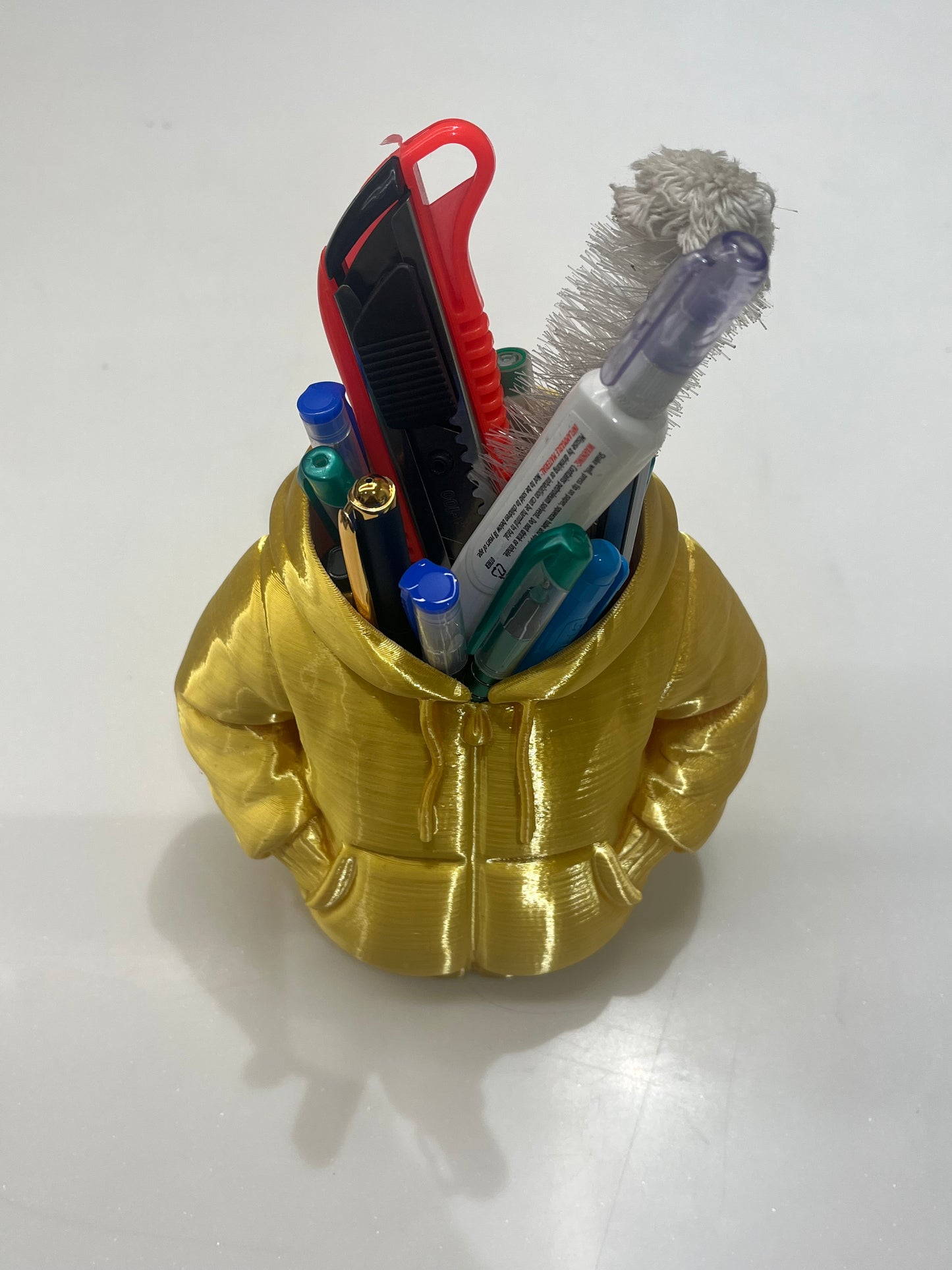 The Mini Hoodie Desk Organizer - 3D Printed Pen Holder is a gold hoodie-shaped desk accessory with hand-in-pocket design, displayed on a white surface and filled with pens, markers, a box cutter, and a cleaning brush.