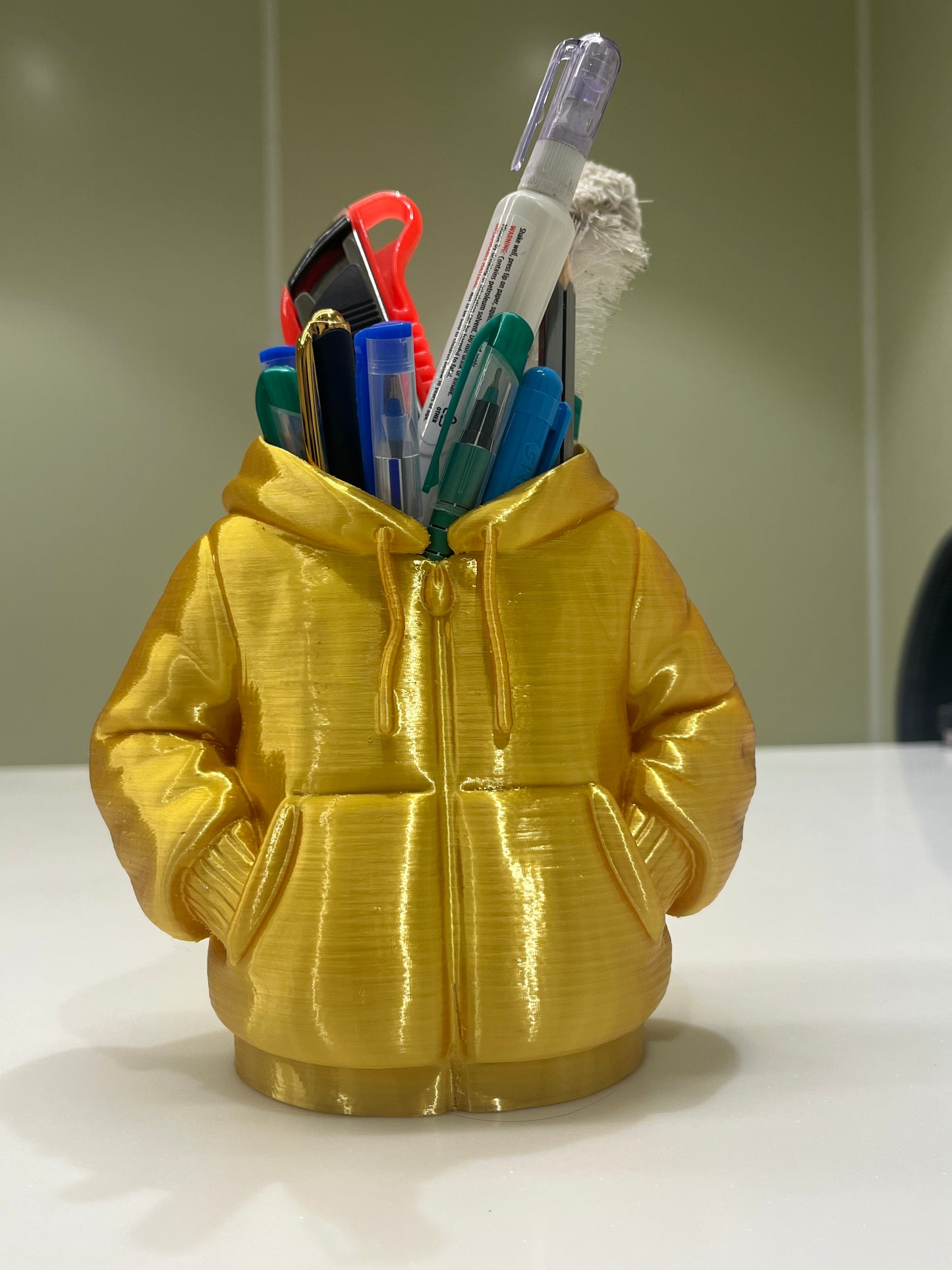 The Mini Hoodie Desk Organizer - 3D Printed Pen Holder in gold holds pens, markers, and a box cutter on a white surface with a light background—a unique and creative desk accessory.