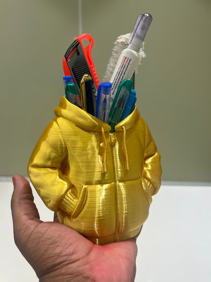 A hand holds the Mini Hoodie Desk Organizer - 3D Printed Pen Holder, a creative yellow desk accessory shaped like a hoodie and filled with pens, a toothbrush, and stationery. The background is plain and blurred.