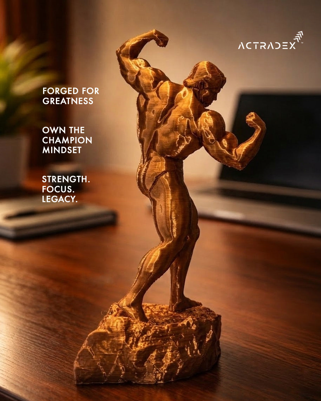 The Arnold Gym Champion - 8” Sculpture features a metallic bronze bodybuilder flexing on a wooden desk. Ideal as an Arnold-inspired desk piece or gym lover’s gift. Text: FORGED FOR GREATNESS. OWN THE CHAMPION MINDSET. Actradex logo top right.